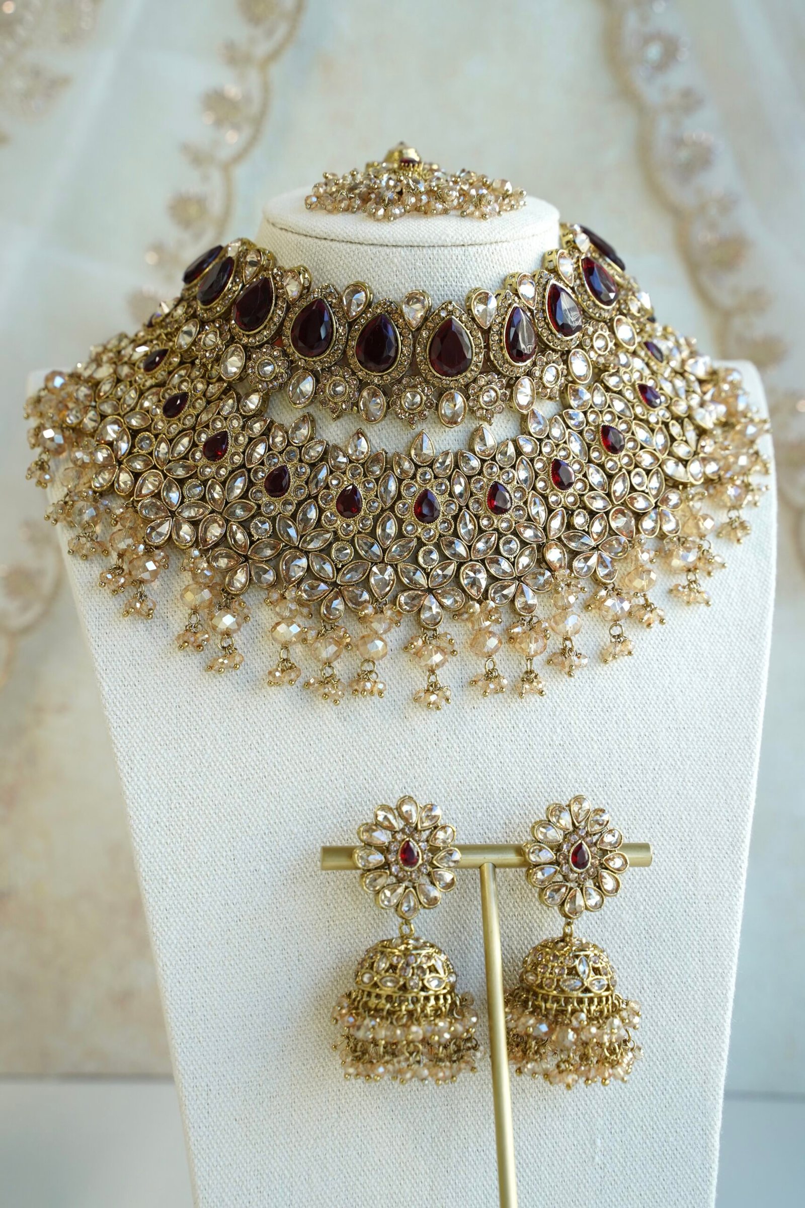Seyora two-layer South Asian bridal necklace set featuring maroon stone and crystal bead settings with a choker, longer necklace, matching jhumka earrings, and a maang tikka