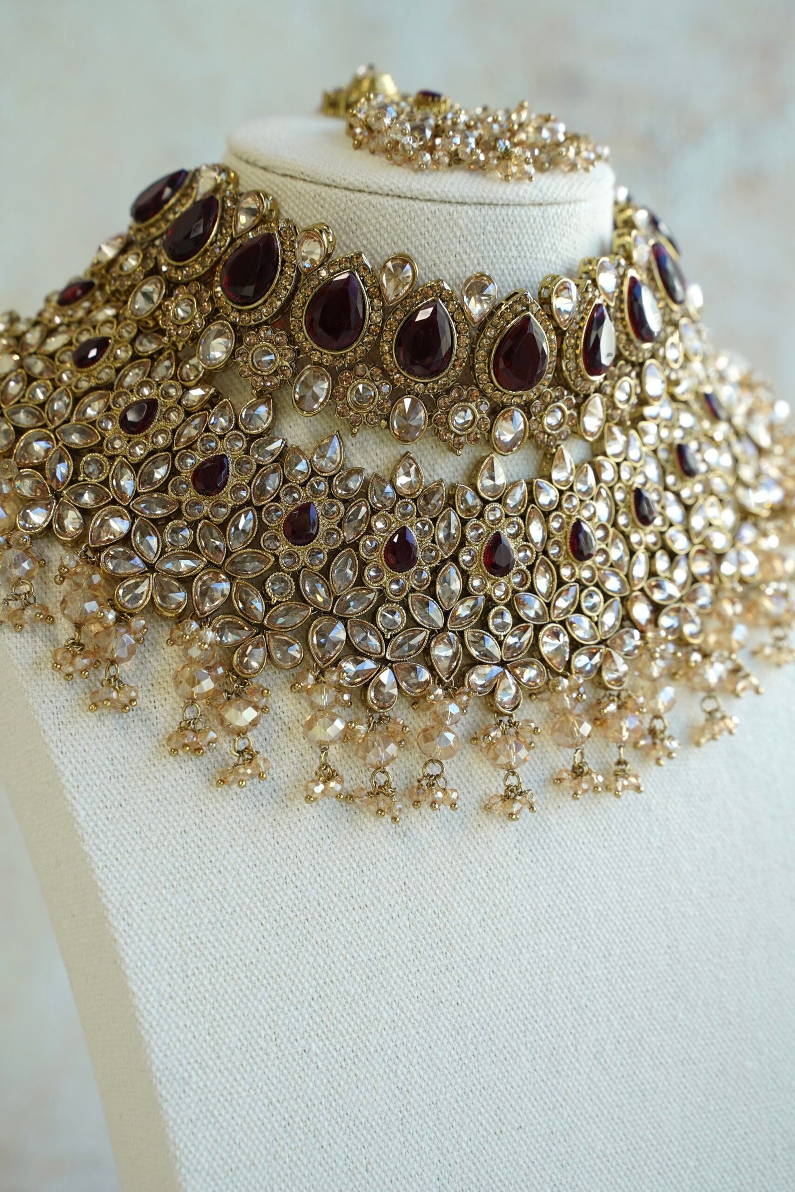 Seyora two-layer South Asian bridal necklace set featuring maroon stone and crystal bead settings with a choker, longer necklace, matching jhumka earrings, and a maang tikka